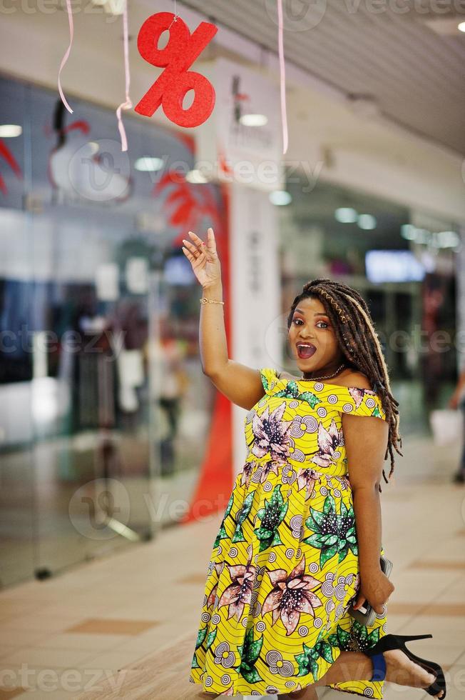 süßes afroamerikanisches mädchen mit kleiner höhe und dreadlocks, trägt ein farbiges gelbes kleid und zeigt den finger auf dem rabattschild im einkaufszentrum. foto