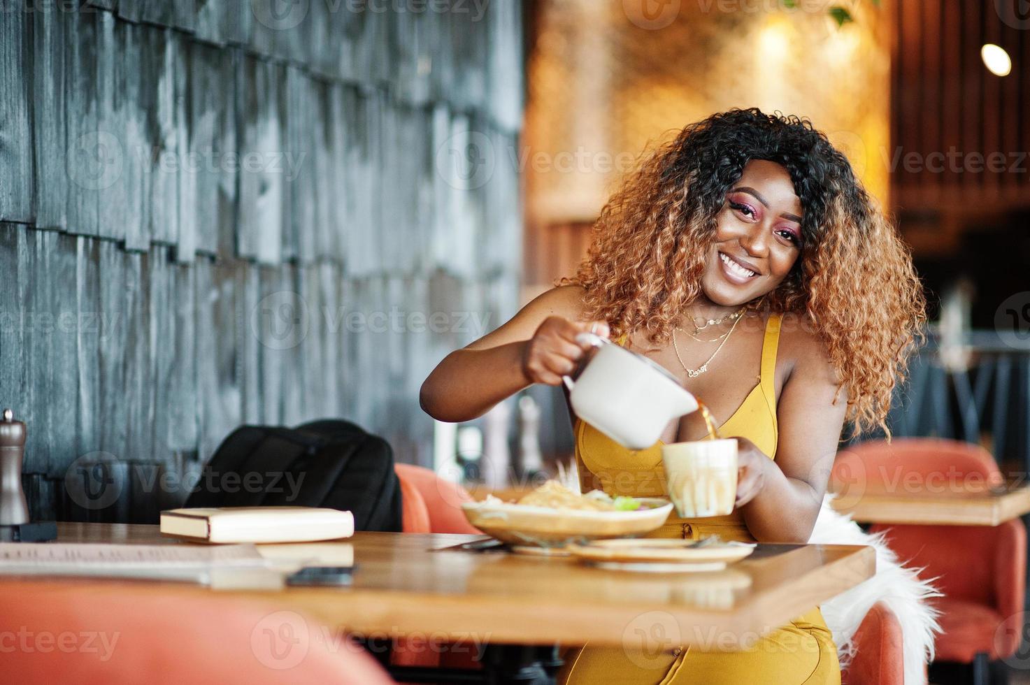 Glamour-Afroamerikanerin im gelben Kleid sitzt im Restaurant und gießt Tee aus der Teekanne. foto