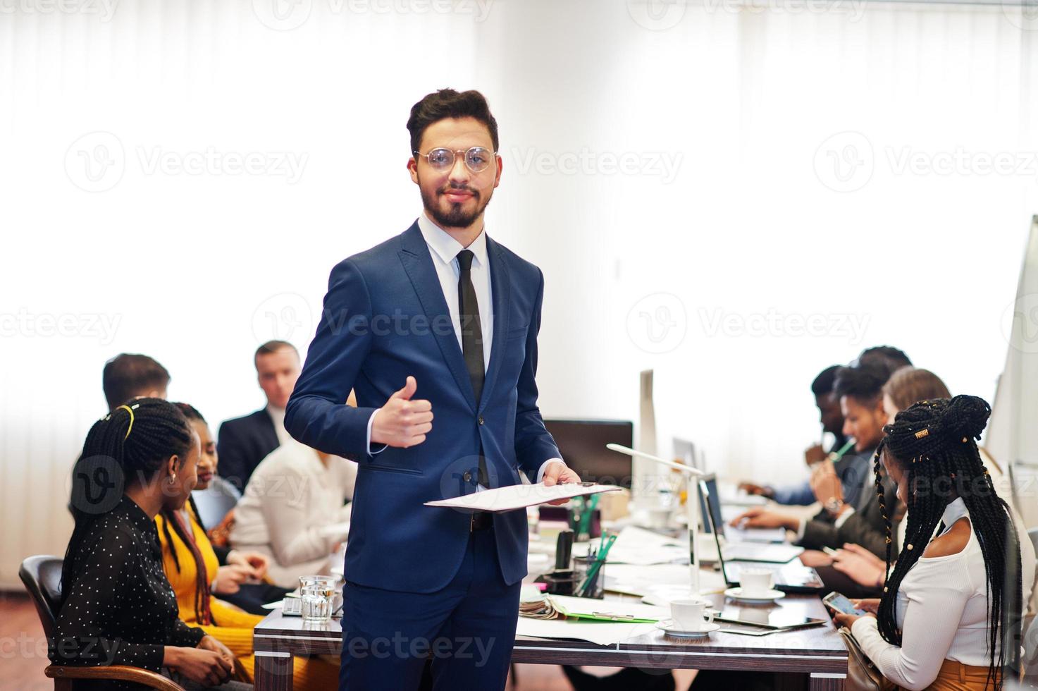 Gesicht eines gutaussehenden arabischen Geschäftsmannes, der eine Zwischenablage hält und den Daumen nach oben auf dem Hintergrund des gemischtrassigen Teamtreffens der Geschäftsleute zeigt und am Bürotisch sitzt. foto