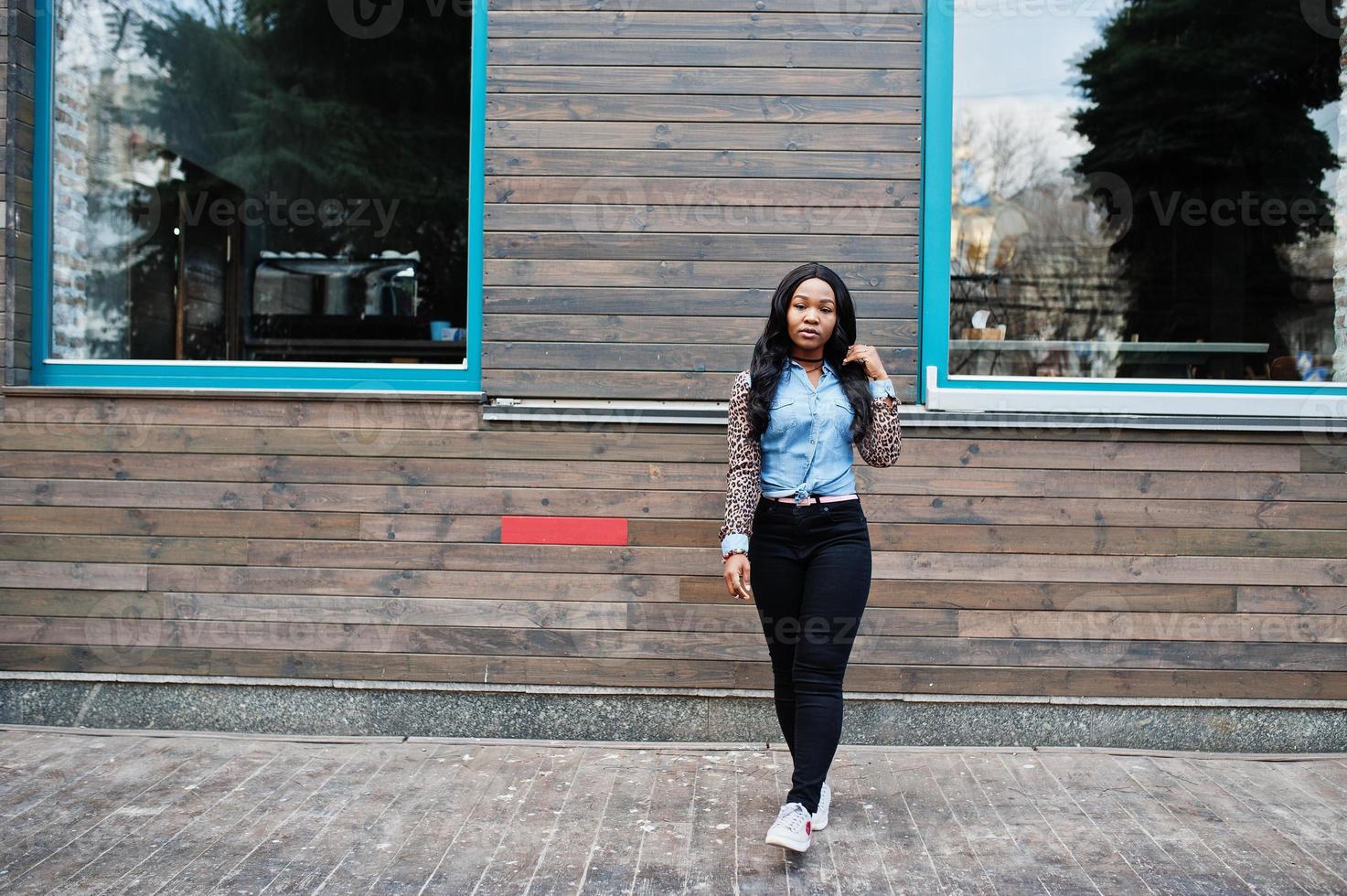 Hipster afroamerikanisches Mädchen mit Jeanshemd und Leopardenärmeln posiert auf der Straße gegen Holzhaus mit Fenstern. foto