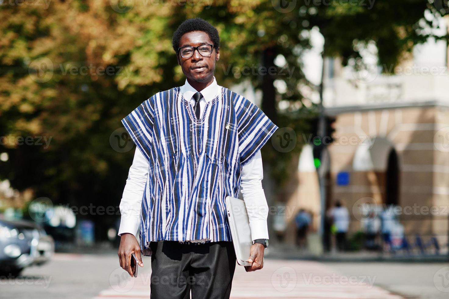 afrikanischer mann in traditioneller kleidung und brille, der mit handy und laptop am zebrastreifen geht. afrika geschäft. foto