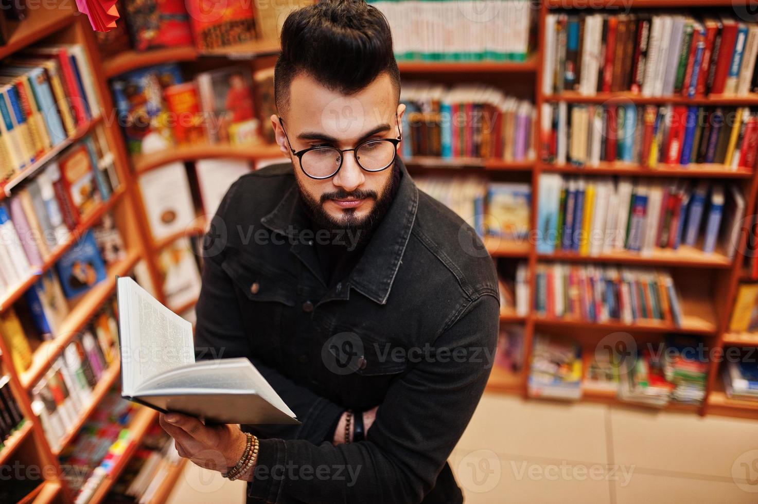 großer, intelligenter arabischer student, trägt schwarze jeansjacke und brille, in der bibliothek mit buch zur hand. foto