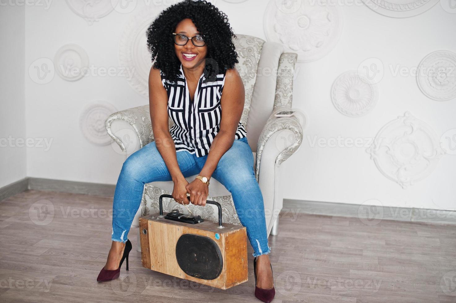 Schöne afroamerikanische Frau mit lockigem Afro-Haar und Brille posierte im Zimmer mit Retro-Tonbandgerät. foto