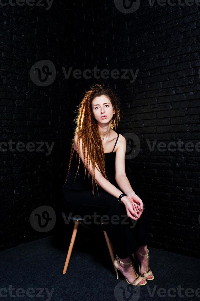Studioaufnahme eines Mädchens in Schwarz auf einem Stuhl mit Dreadlocks auf Backsteinhintergrund. foto