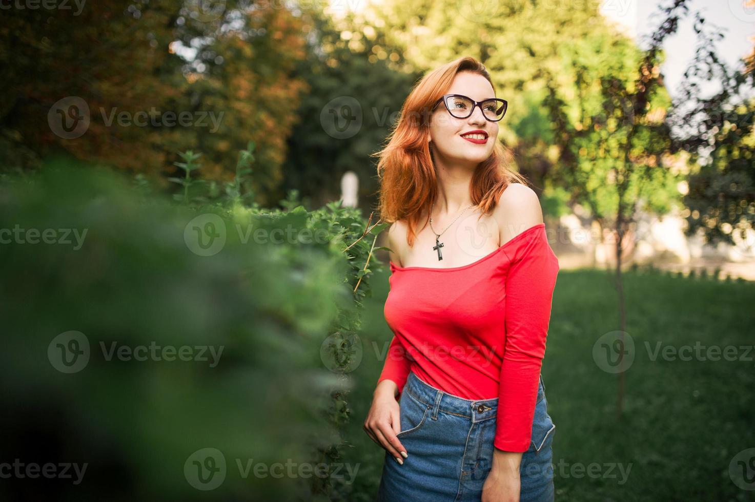 attraktive rothaarige frau mit brille, tragen auf roter bluse und jeansrock, die im grünen park posieren. foto