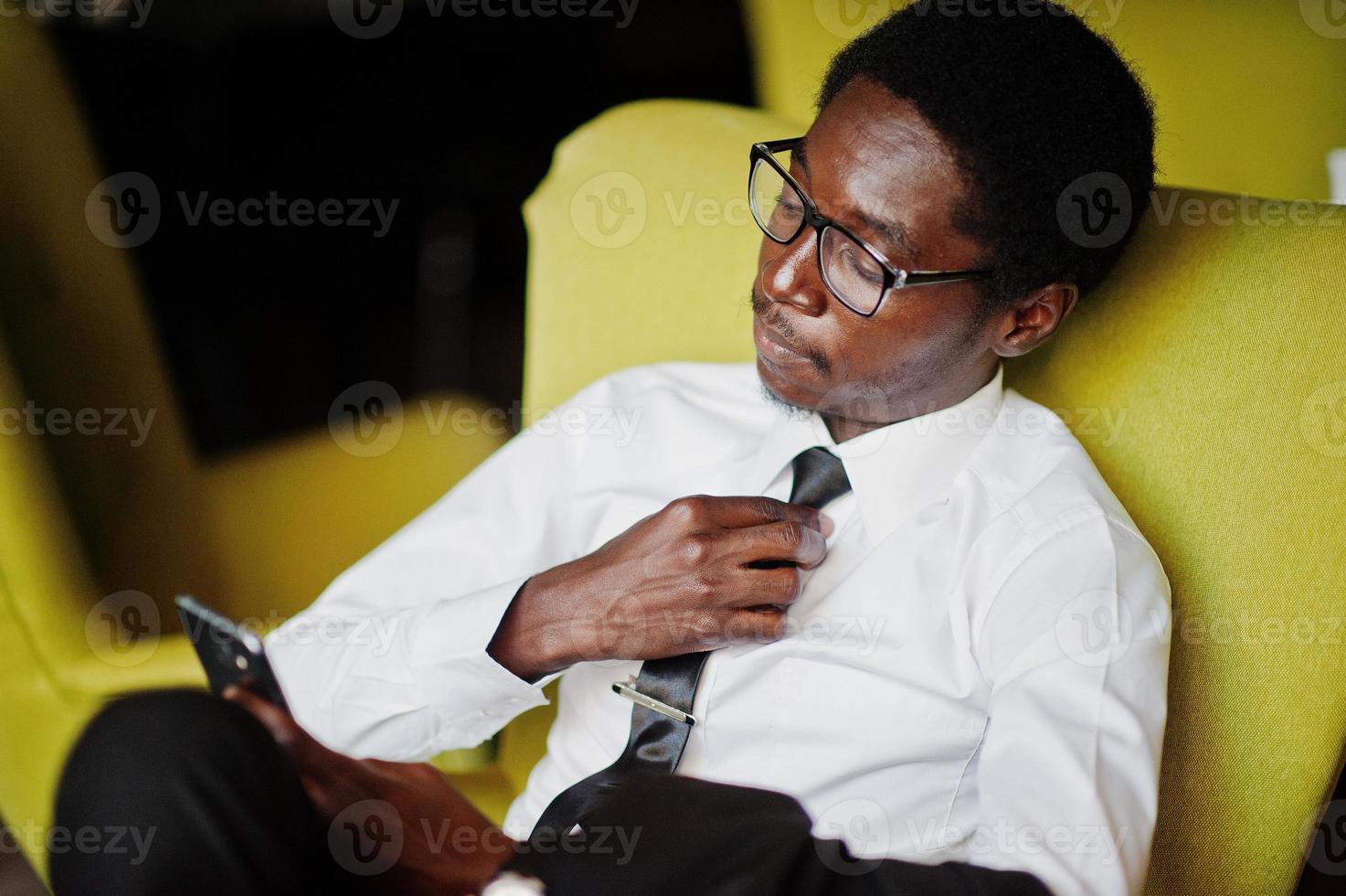 business afroamerikaner tragen weißes hemd, krawatte und brille im büro, sitzen auf stuhl und halten handy in der hand. foto