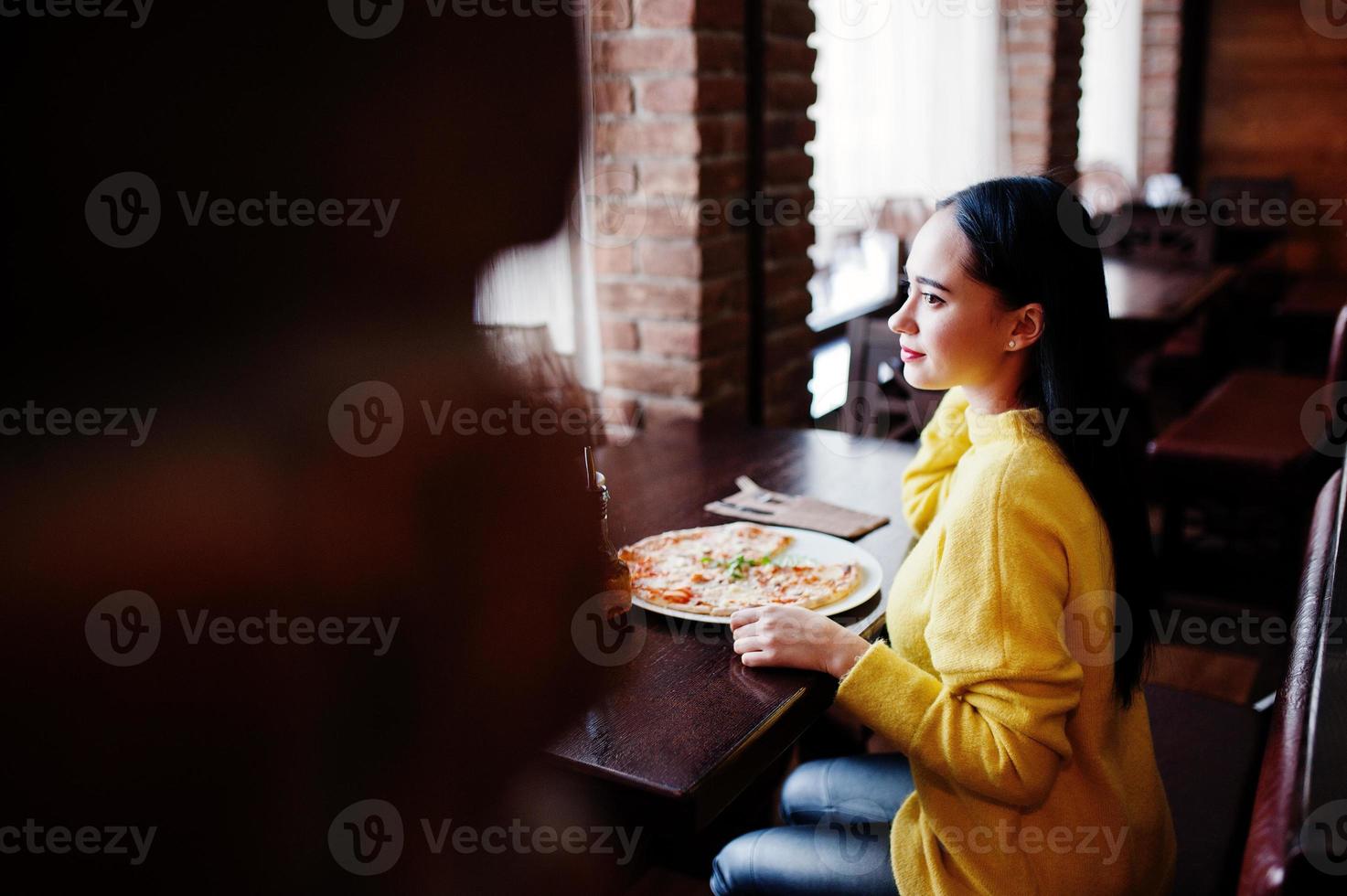 lustiges brünettes mädchen im gelben pullover, der pizza im restaurant isst. foto