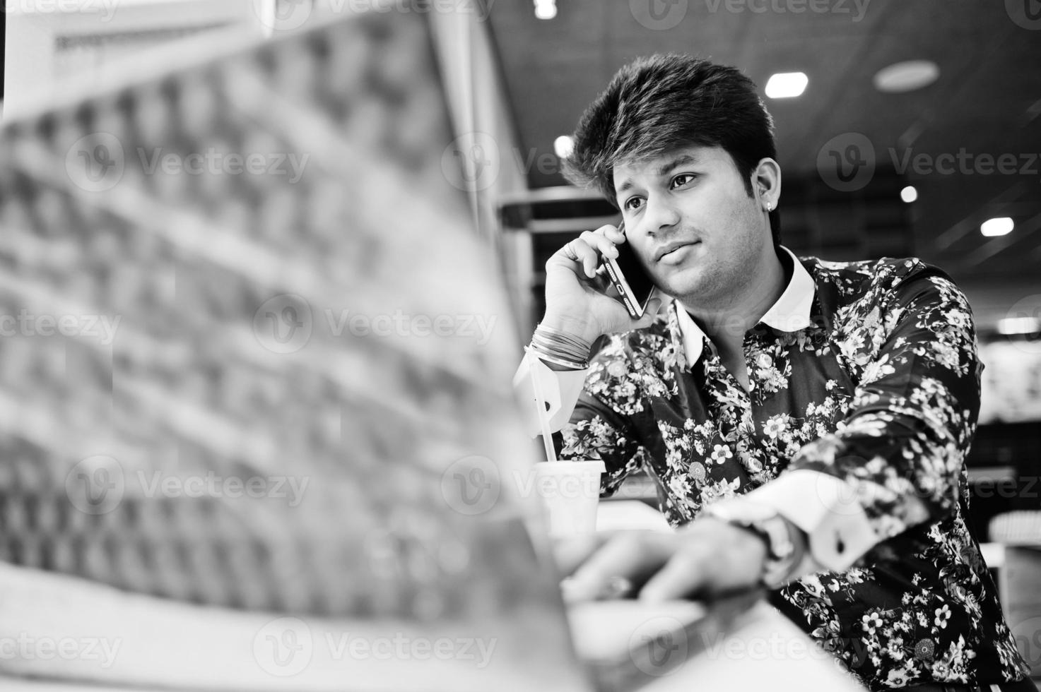 junger männlicher indischer freiberufler, der auf offenem laptop in einem fast-food-café sitzt, hübscher asiatischer mann, der während der arbeit am net-book in einem gemütlichen café telefoniert. foto