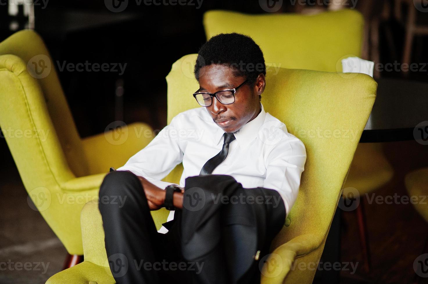 business afroamerikaner trägt weißes hemd, krawatte und brille im büro, sitzt auf stuhl und schaut auf uhren. foto