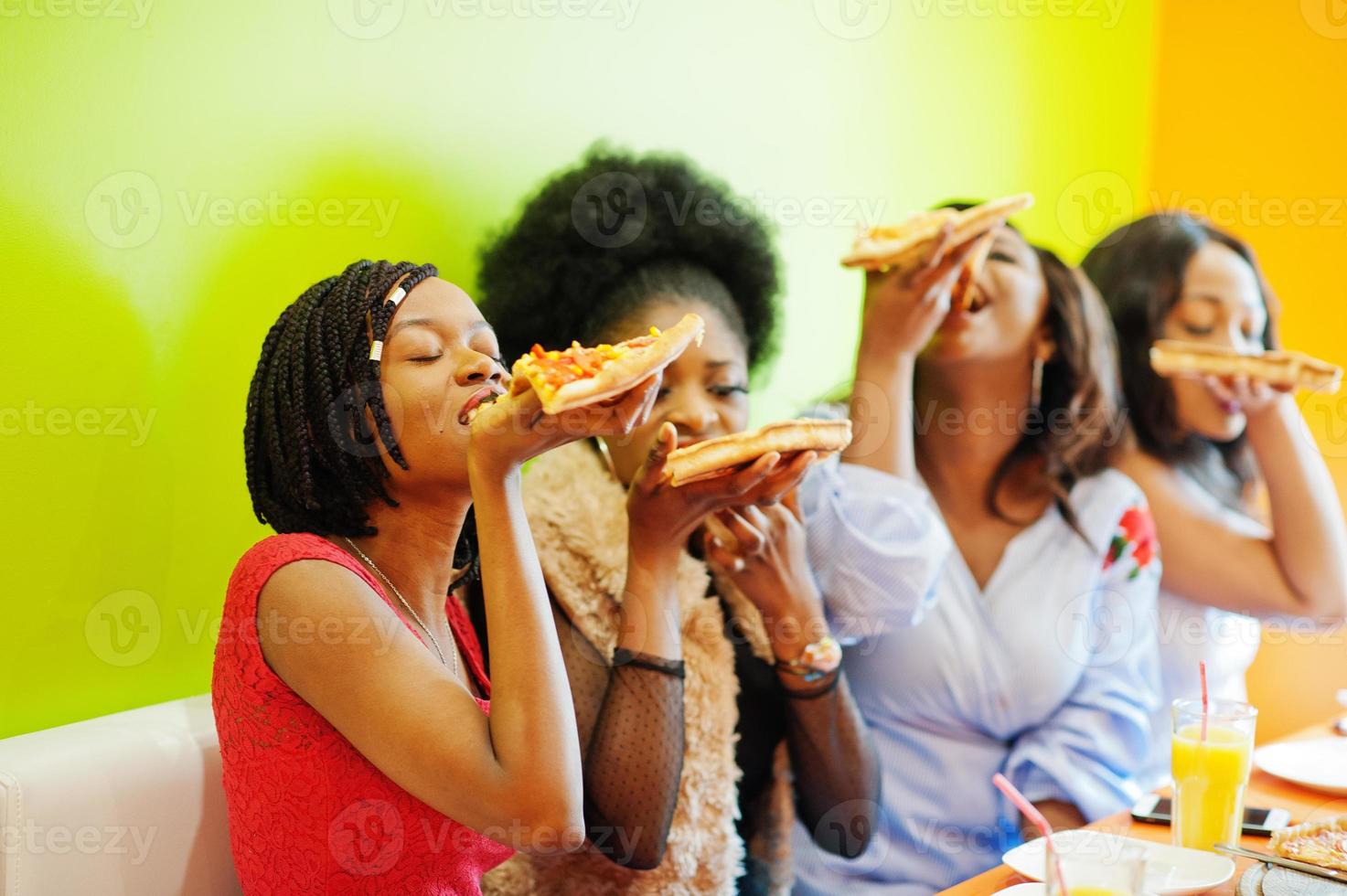 Vier junge afrikanische Mädchen in einem bunten Restaurant essen Pizzastücke in den Händen. foto