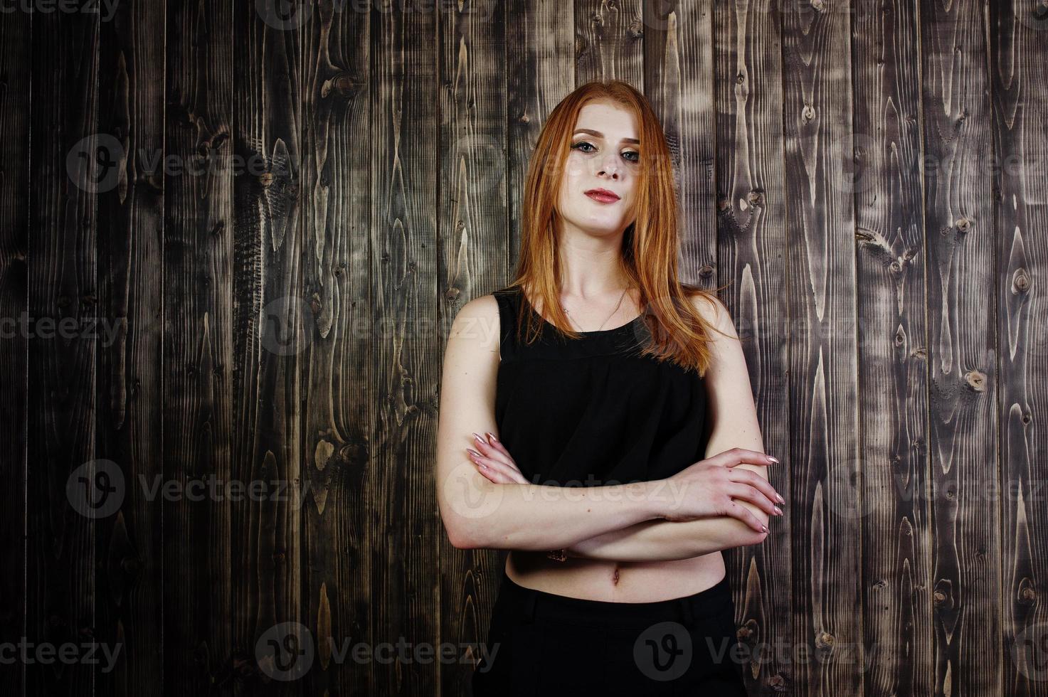 Porträt eines schönen rothaarigen Mädchens in schwarzem Oberteil und schwarzem Rock, das im Studio neben der Holzwand posiert. foto