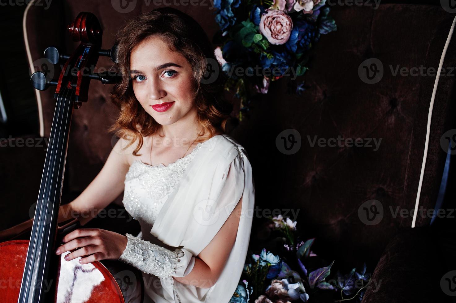 hübscher junger Mädchenmusiker im weißen Kleid mit Kontrabass, der auf braunem Vintage-Sofa sitzt. foto