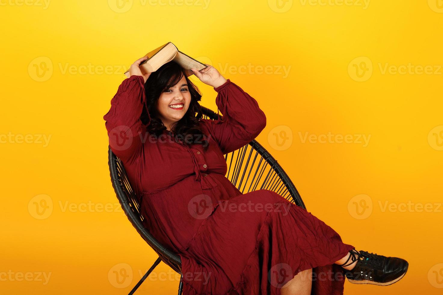 attraktive südasiatische frau in tiefrotem kleiderkleid posierte im studio auf gelbem hintergrund sitzend auf stuhl mit buch. foto