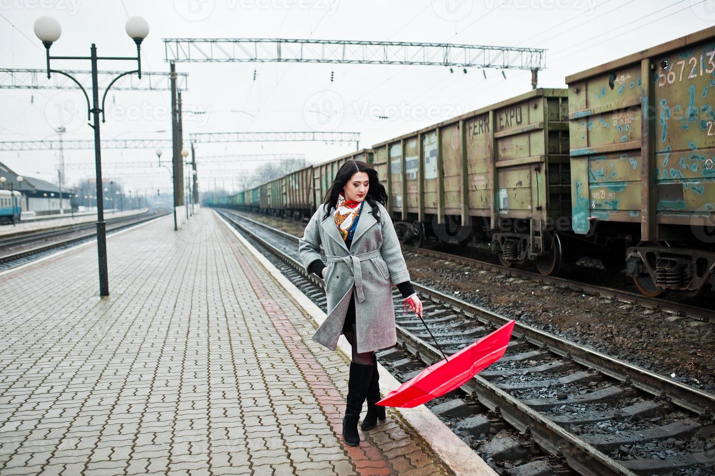 Brünettes Mädchen im grauen Mantel mit rotem Regenschirm im Bahnhof. foto