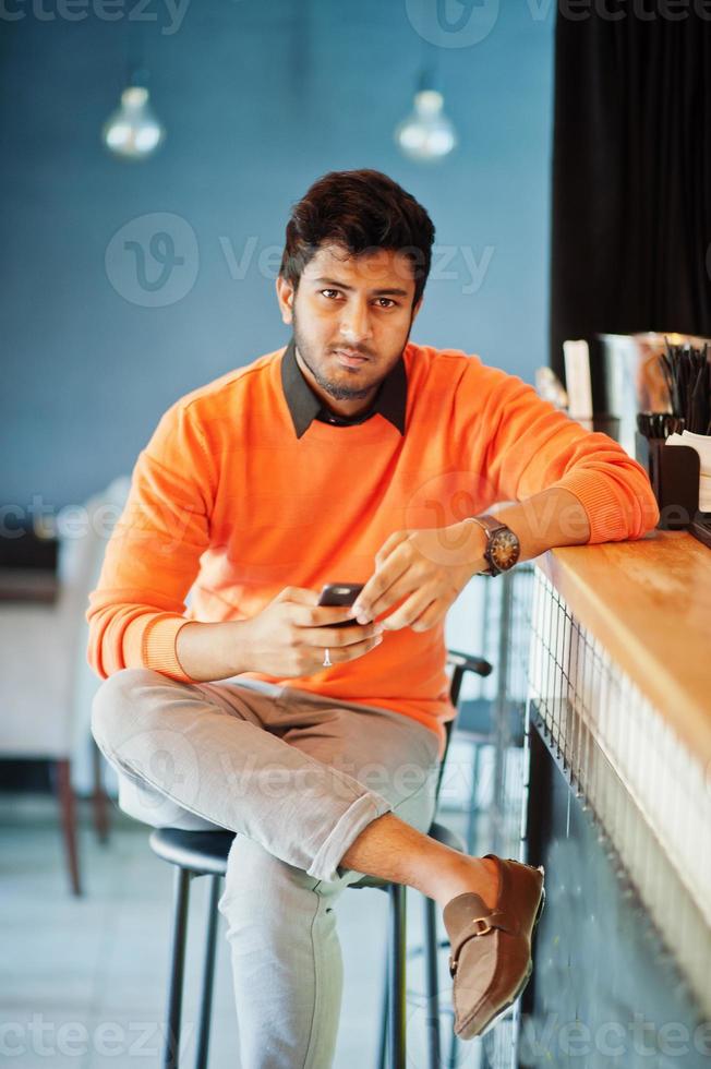 Selbstbewusster junger indischer Mann in orangefarbenem Pullover sitzt an der Theke im Café mit Handy zur Hand. foto