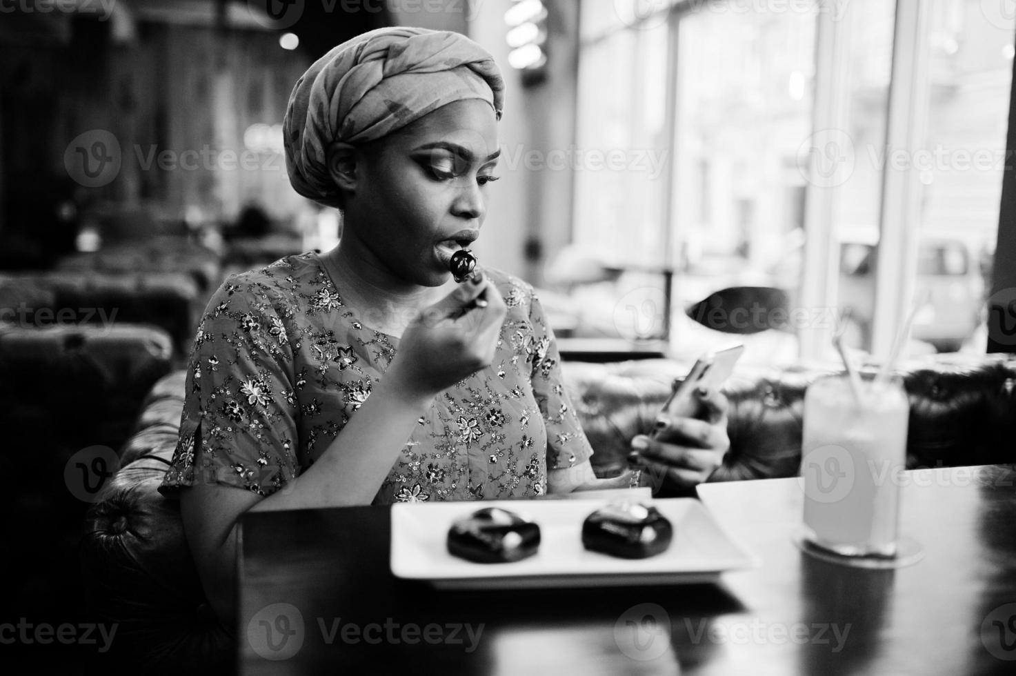 stilvolle afrikanische frau in rotem hemd und hut posierte innencafé, iss schokoladendessertkuchen und schaut aufs handy. foto