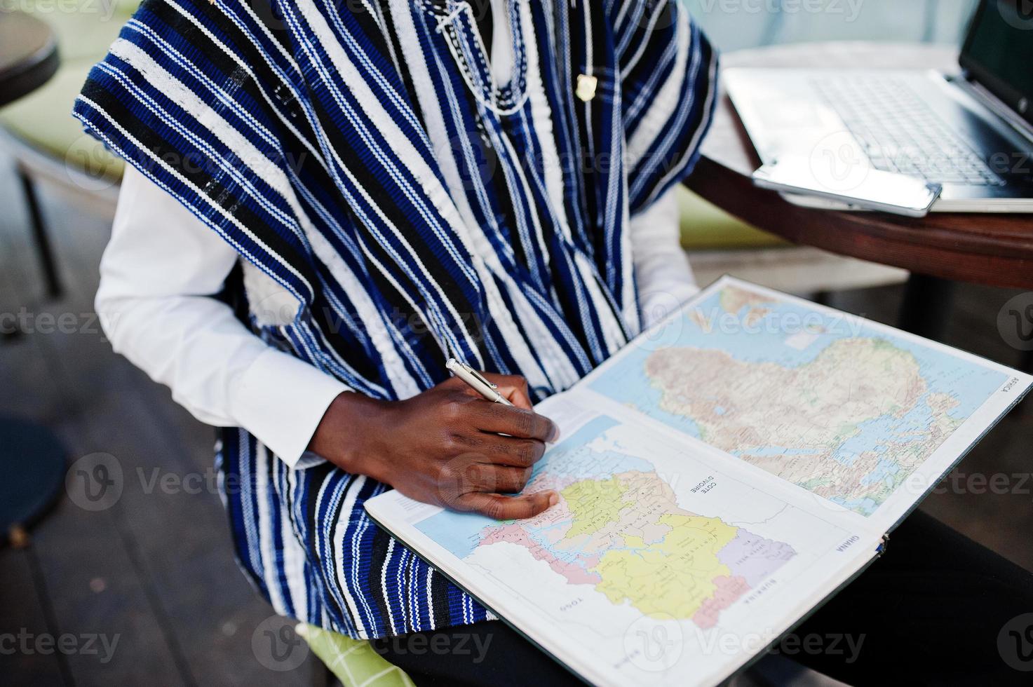 afrikanischer Mann in traditioneller Kleidung, der hinter einem Laptop im Café im Freien sitzt und auf der Karte von Afrika und Ghana auf sein Notizbuch blickt. foto