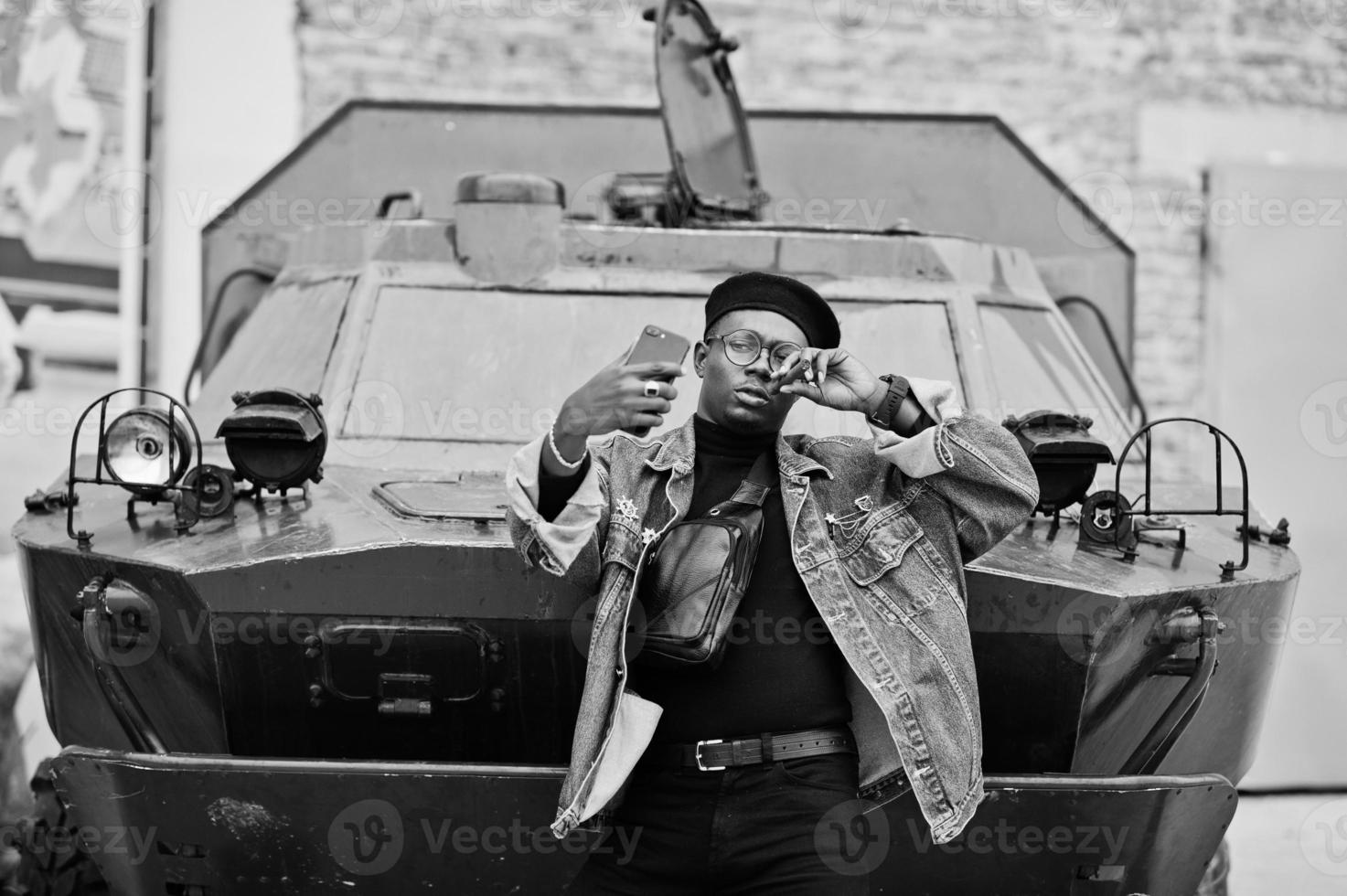 afroamerikanischer mann in jeansjacke, baskenmütze und brille, mit zigarre, die gegen ein gepanzertes btr-militärfahrzeug posiert und selfie auf dem handy macht. foto