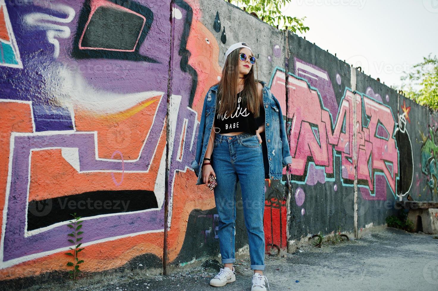 stilvolles lässiges hipster-mädchen in mütze, sonnenbrille und jeanskleidung, musik hören über kopfhörer des handys gegen große graffitiwand. foto