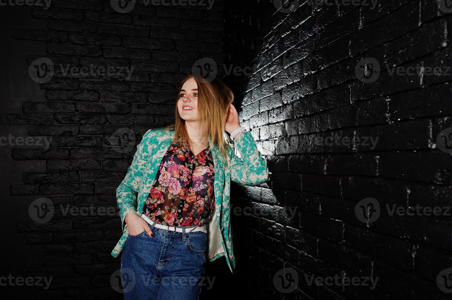 stylisches blondes Mädchen in Jacke und Jeans gegen schwarze Ziegelwand im Studio. foto