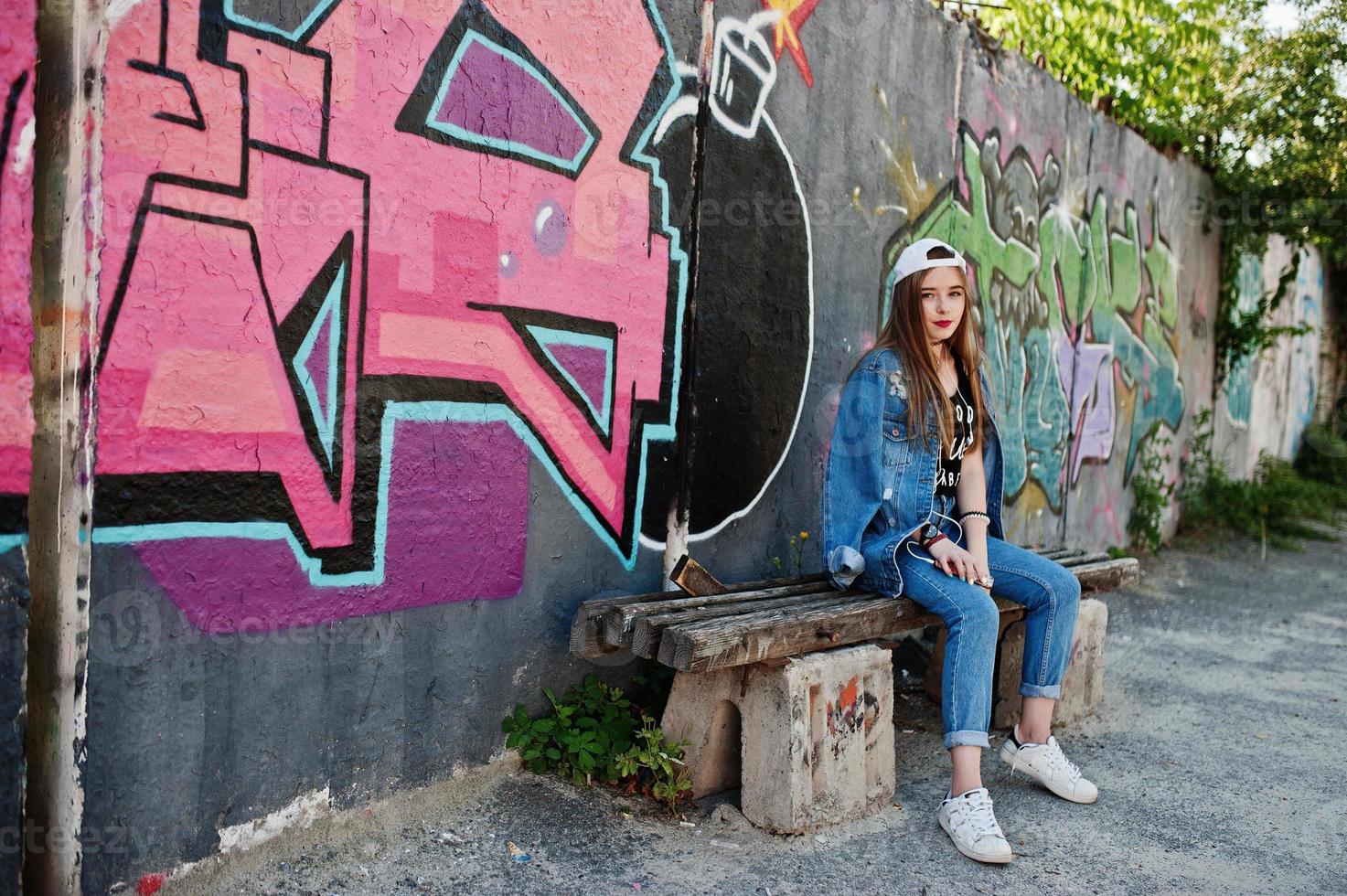 stilvolles, lässiges hipster-mädchen in mütze und jeans, das musik über kopfhörer des handys gegen große graffitiwand mit bombe hört. foto