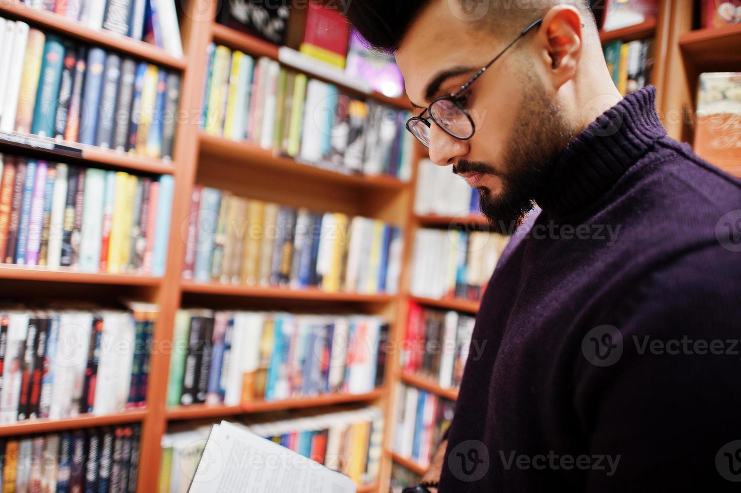 großer, intelligenter arabischer student, trägt einen violetten rollkragenpullover und eine brille, liest in der bibliothek ein buch. foto