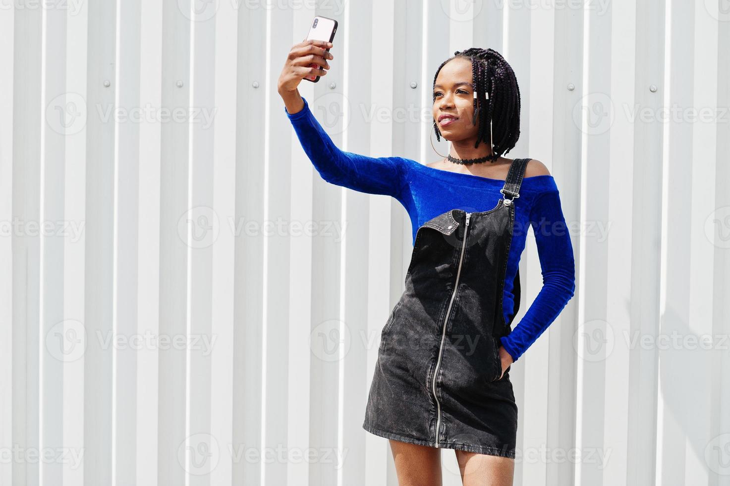 afrikanische frau in jeansoverallrock und blauem kleid posierte vor weißer stahlwand und machte selfie am telefon. foto