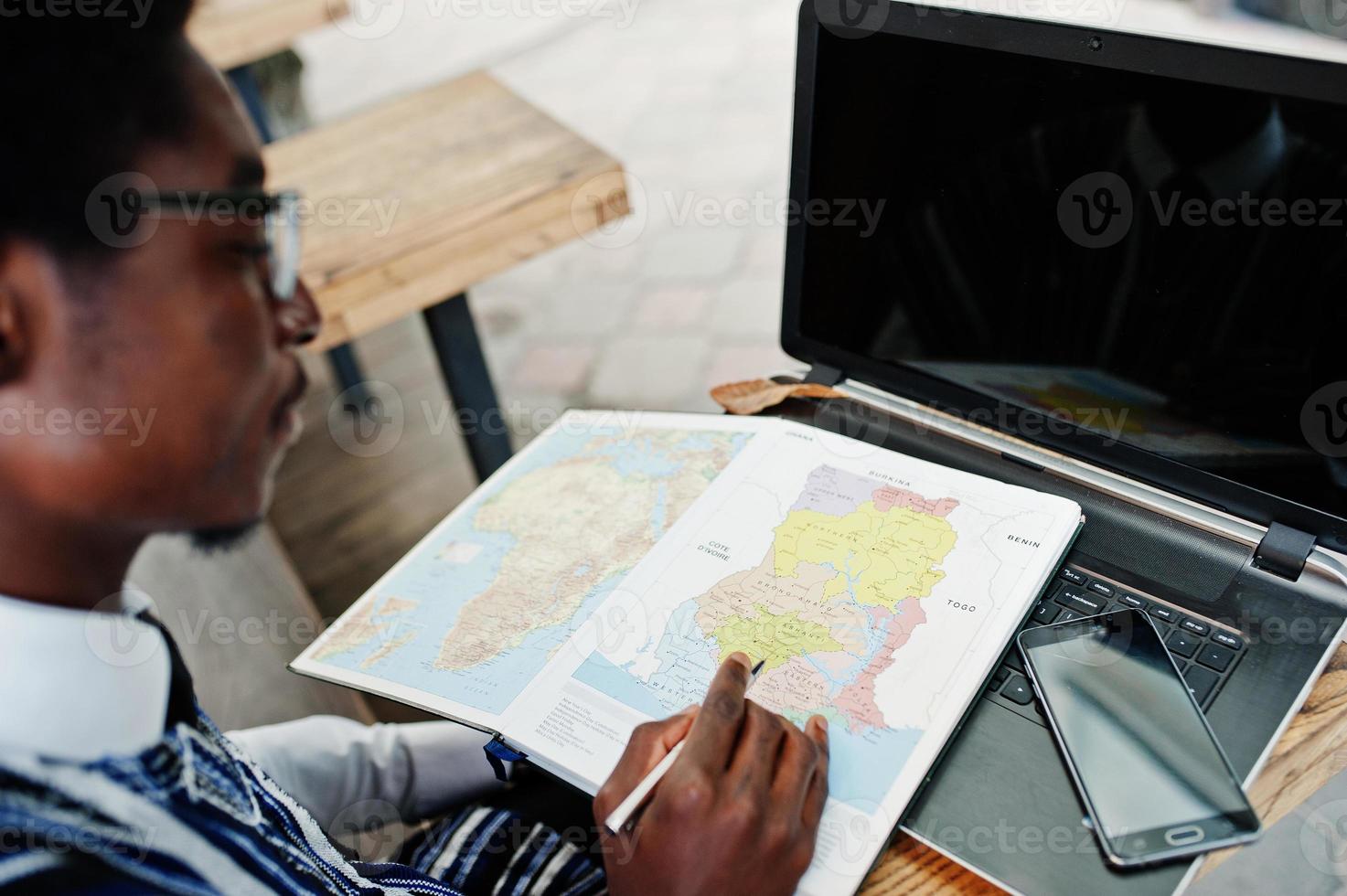 afrikanischer Mann in traditioneller Kleidung und Brille sitzt hinter Laptop im Café im Freien und schaut auf der Karte von Afrika und Ghana auf sein Notizbuch. foto