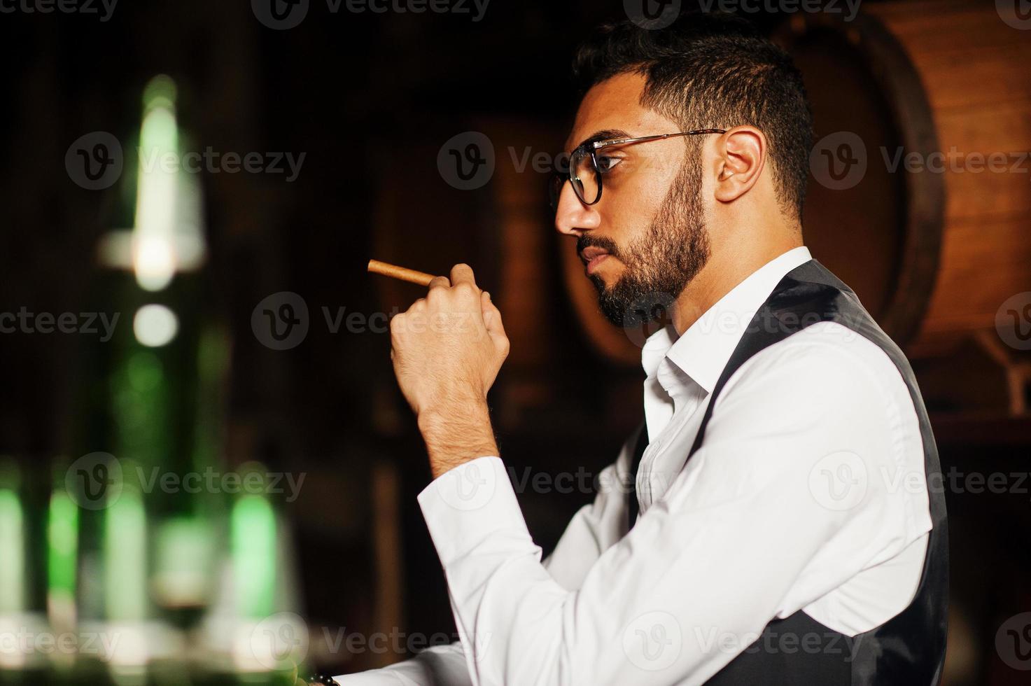 hübscher, gut gekleideter arabischer mann mit einem glas whisky und einer zigarre, die im pub posiert. foto