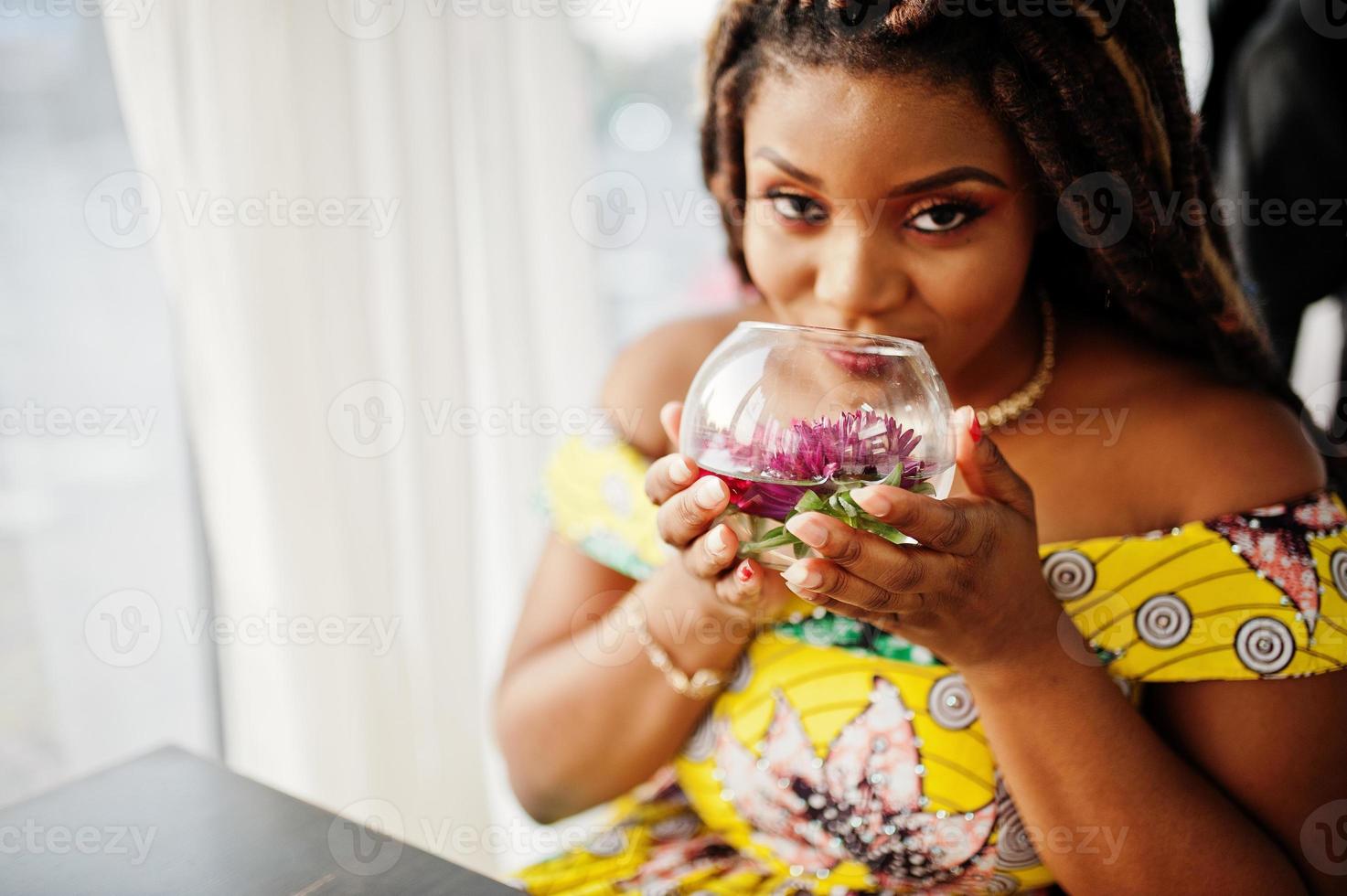 süßes kleines afroamerikanisches mädchen mit dreadlocks, trägt ein farbiges gelbes kleid und sitzt im café mit blume im glas. foto