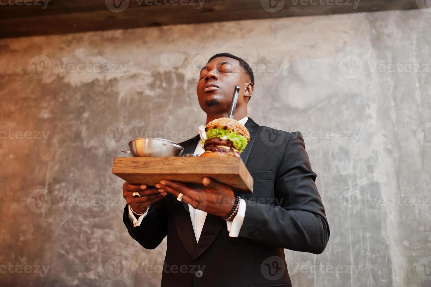 respektabler junger afroamerikaner im schwarzen anzug hält tablett mit doppelburger gegen graue wand. er atmet den Duft eines leckeren Hamburgers. foto