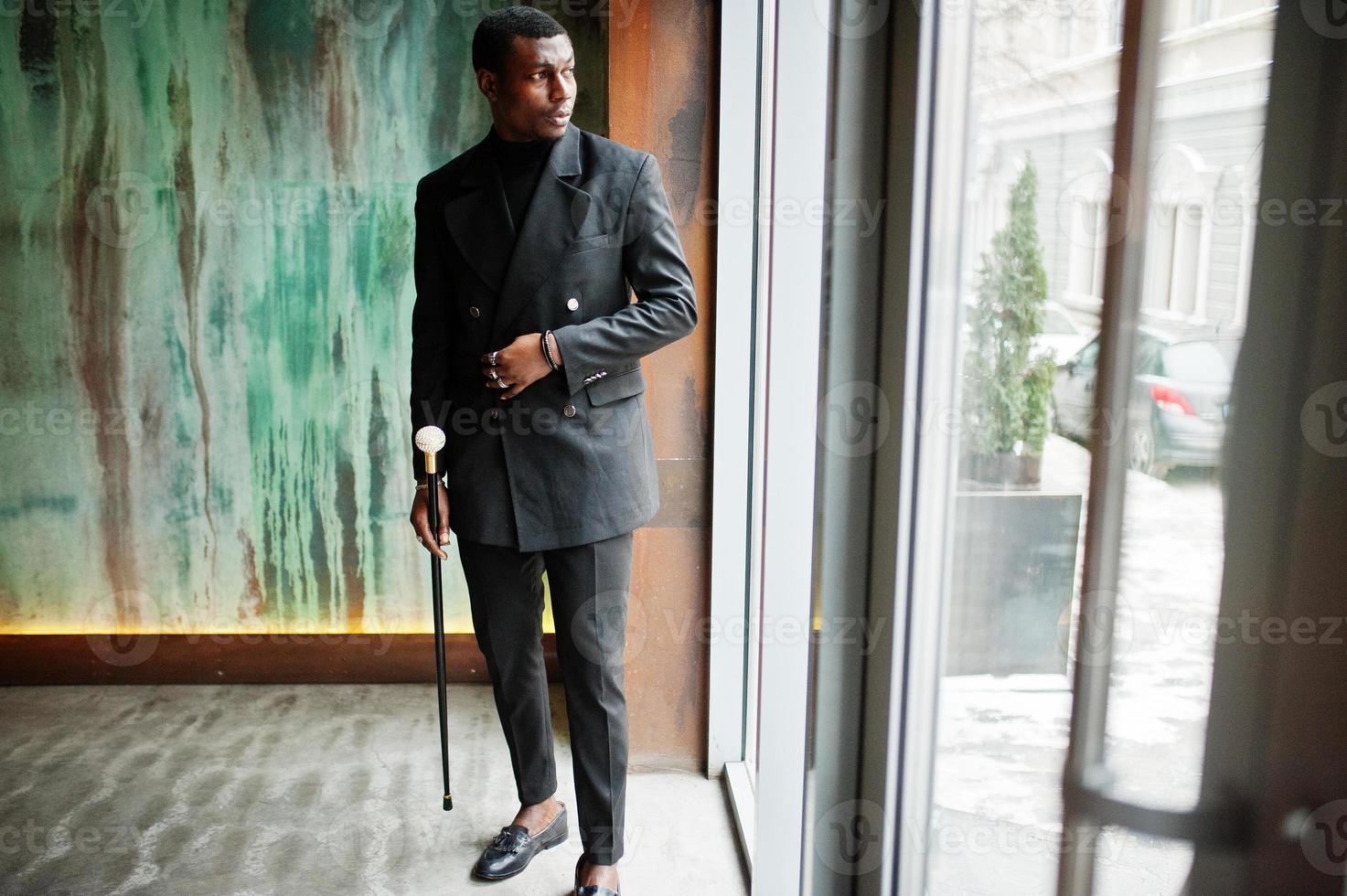 Stilvoller afroamerikanischer Gentleman in eleganter schwarzer Jacke, der einen Retro-Spazierstock als Rohrkolben oder einen Kippstock mit goldenem Diamantkugelgriff hält. reicher modischer Afro-Mann. foto