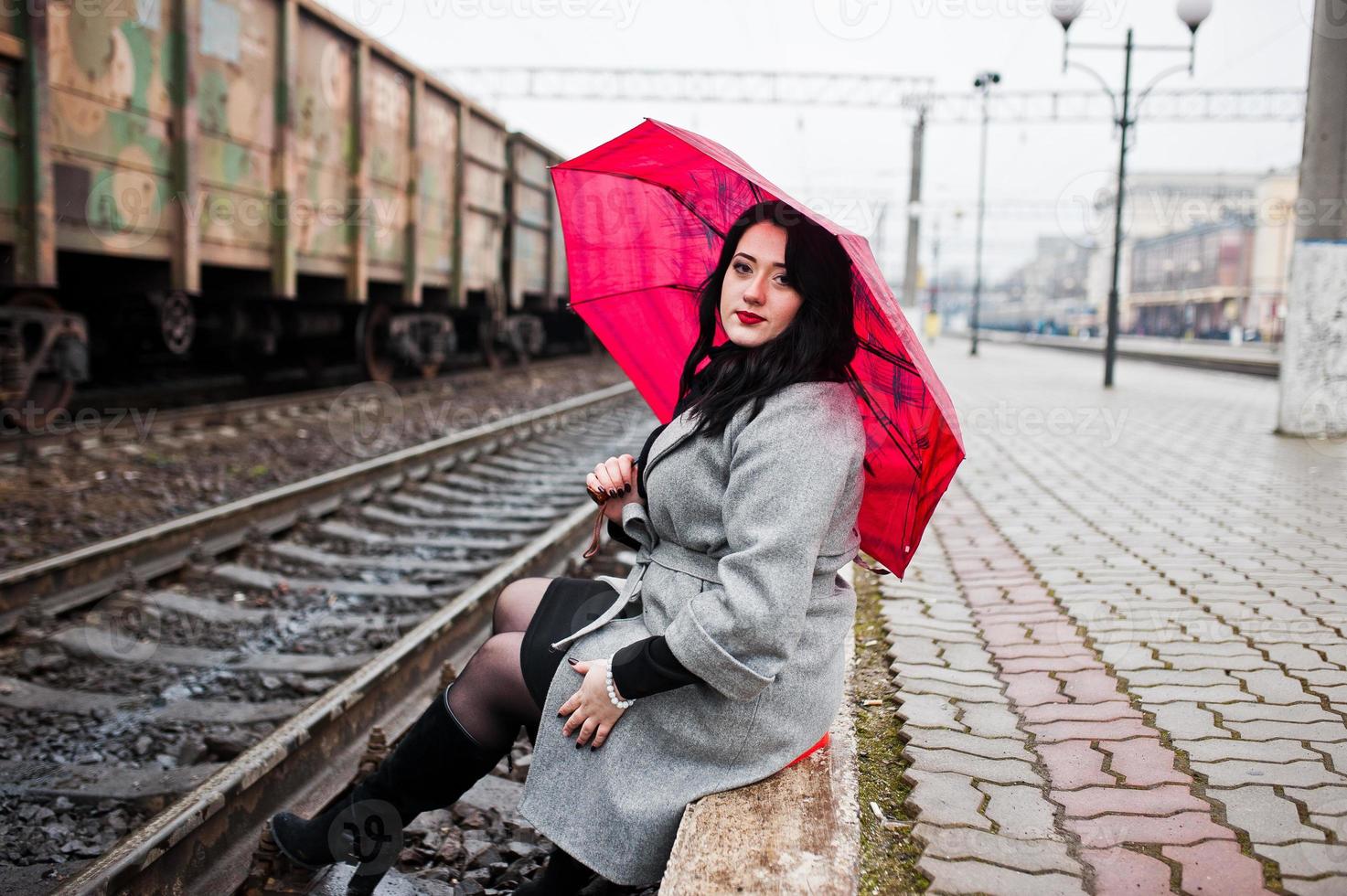 Brünettes Mädchen im grauen Mantel mit rotem Regenschirm im Bahnhof. foto