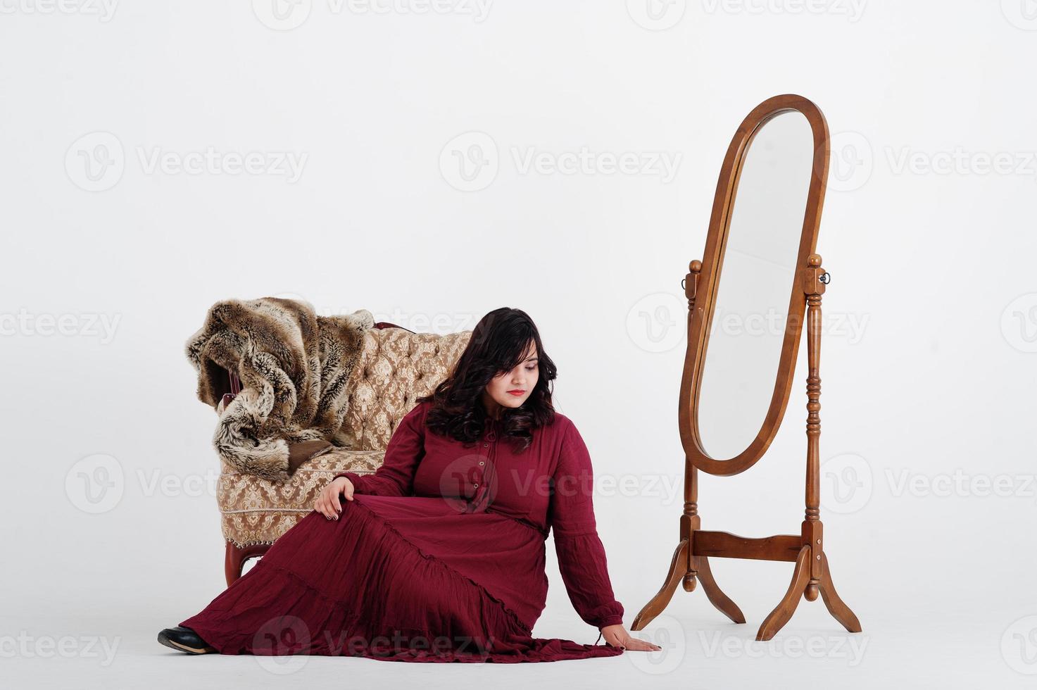 Attraktive südasiatische Frau in tiefrotem Kleid posiert im Studio auf weißem Hintergrund vor Spiegel und Stuhl. foto