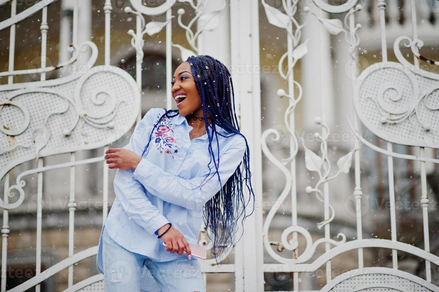 Stilvolles afrikanisch-amerikanisches Mädchen mit Dreadlocks, das Handy zur Hand hält, im Freien vor weißen königlichen Toren bei Schneewetter. foto