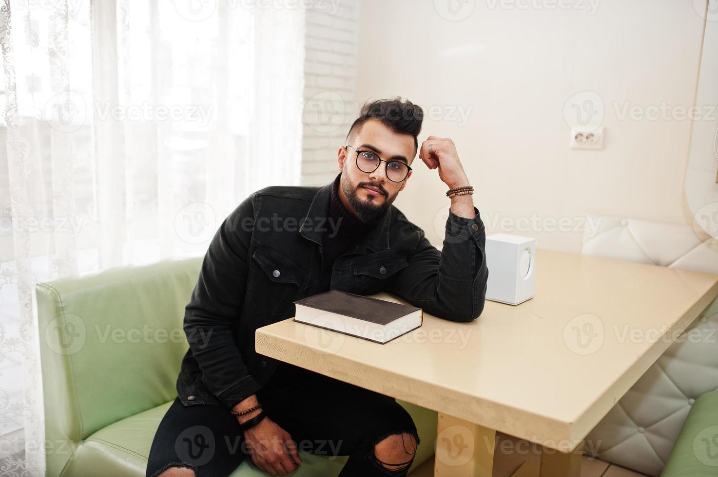 arabischer mann trägt schwarze jeansjacke und brille im café sitzend, buch lesen. stilvoller und modischer arabischer Modelltyp. foto