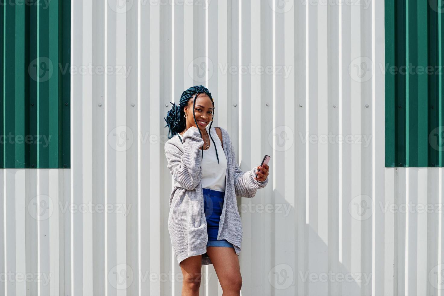 afrikanische frau mit dreadlockshaar, in jeansshorts, die mit handy in der hand vor einer weißen stahlwand posiert. foto