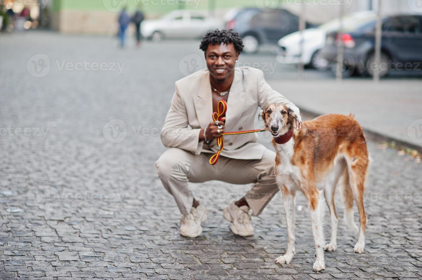 Stilvoller Afro-Mann im beigen Old-School-Anzug mit russischem Barsoi-Hund. Modischer junger afrikanischer Mann in Freizeitjacke auf nacktem Oberkörper. foto