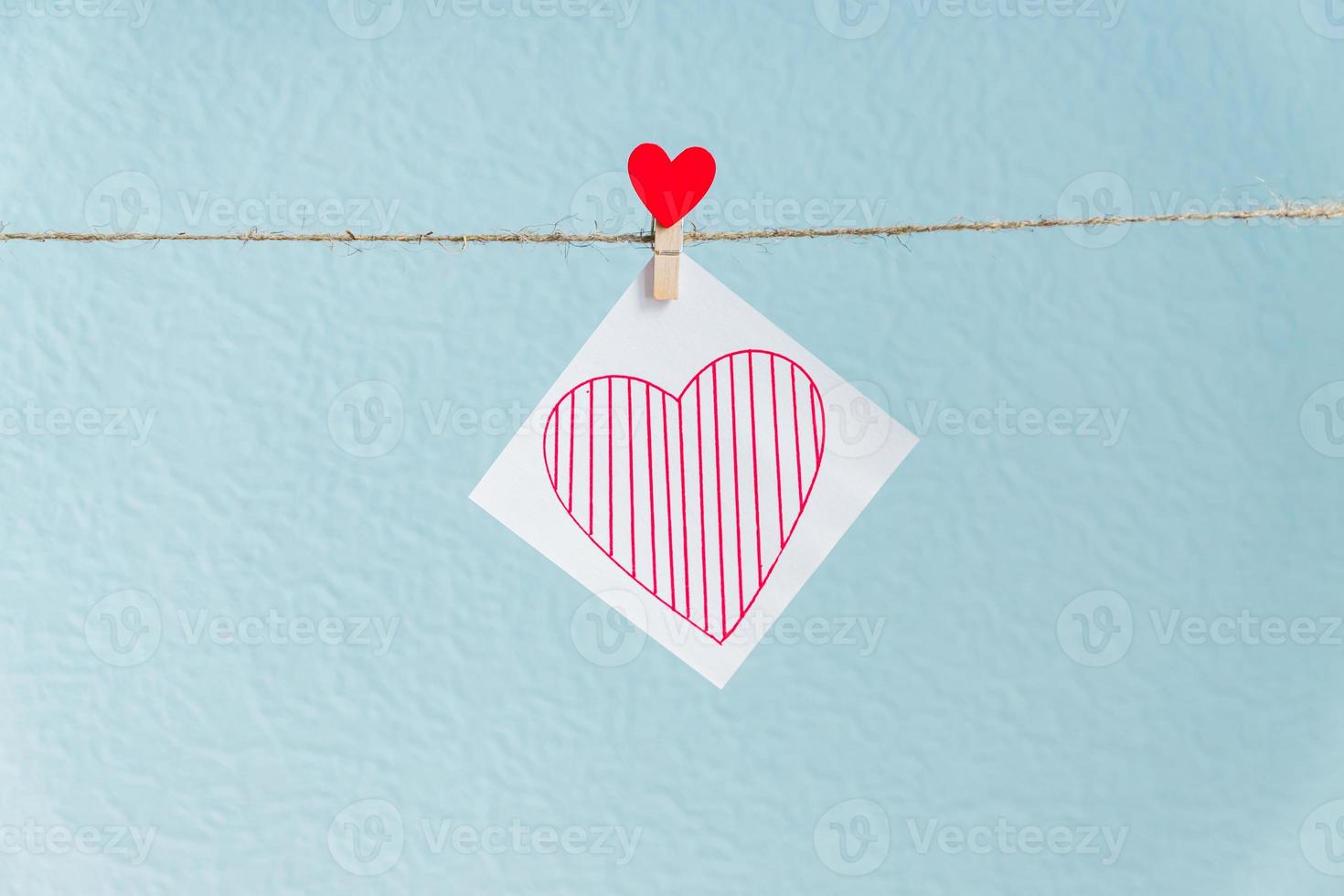 Liebesherzstift des roten Valentinsgrußes, der an der natürlichen Schnur gegen blauen Hintergrund hängt. gezeichnetes Herz auf Papierstück. foto