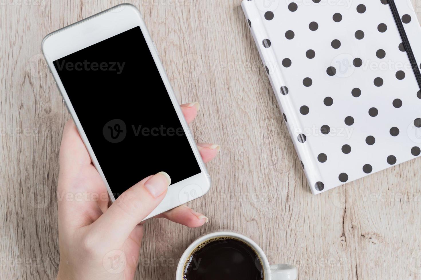 Geschäfts- und Bürokonzept - Frauenhand, die weiße Smartwhone neben dem schwarz-weiß gepunkteten Notizbuch und einer Tasse schwarzen Kaffee auf einem Holztisch hält. Ansicht von oben. foto