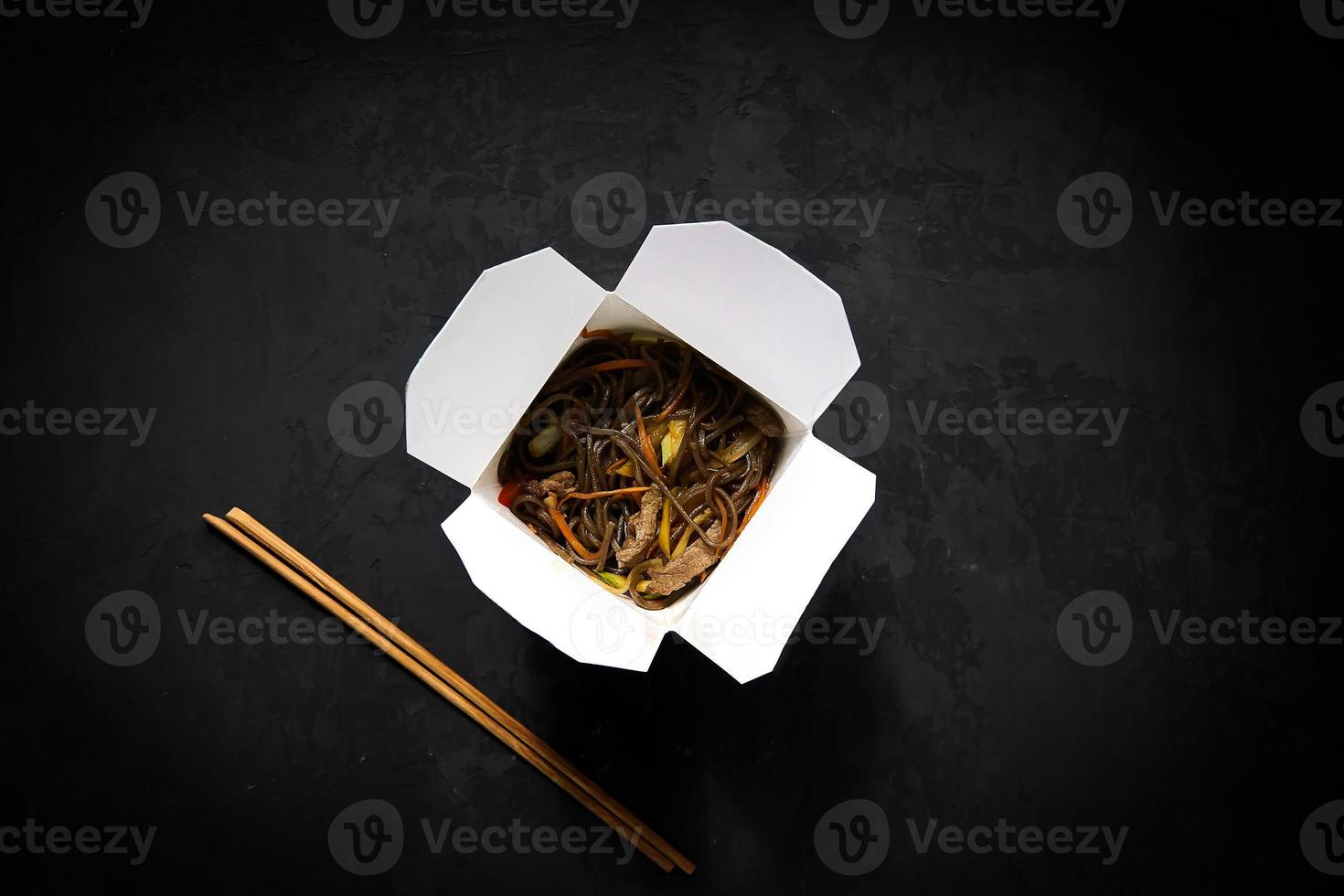 Lieferservice für asiatische Restaurants. Soba-Nudeln mit Fleisch, Gemüse und Sojasauce in weißer Papierbox zum Mitnehmen auf schwarzem Hintergrund mit Essstäbchen, Draufsicht, Kopierraum foto