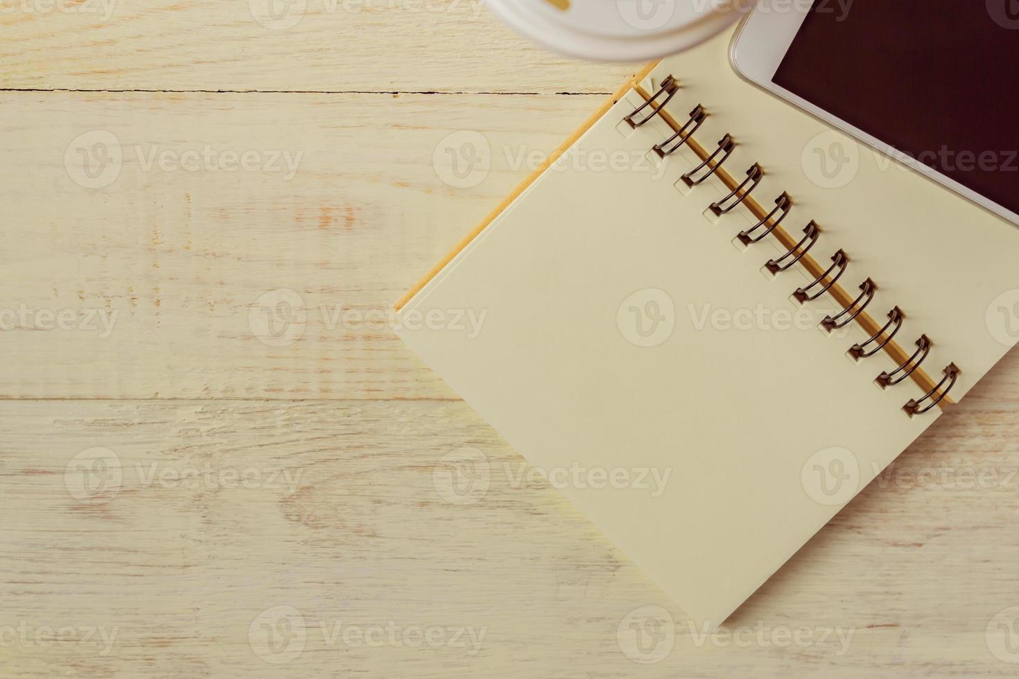 Handwerk Einweg-Kaffeetasse aus Papier, Notizbuch und weißes Smartphone auf Holzhintergrund mit Kopierraum foto