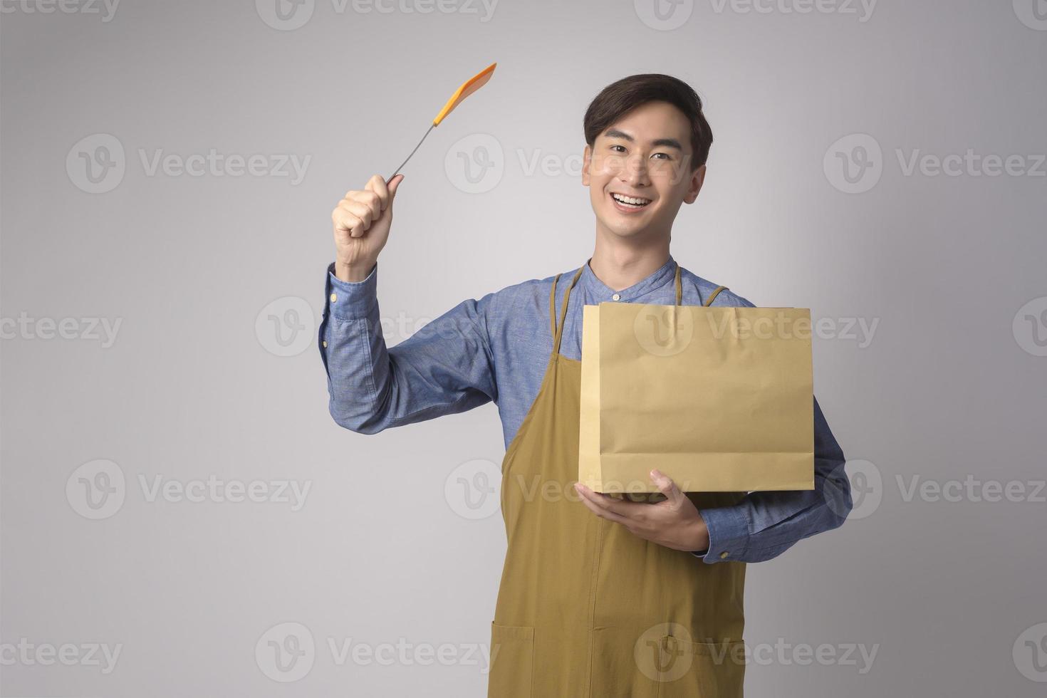 Porträt des jungen asiatischen Mannes mit Schürze mit Papiertüte und Turner auf weißem Hintergrund Studio. foto