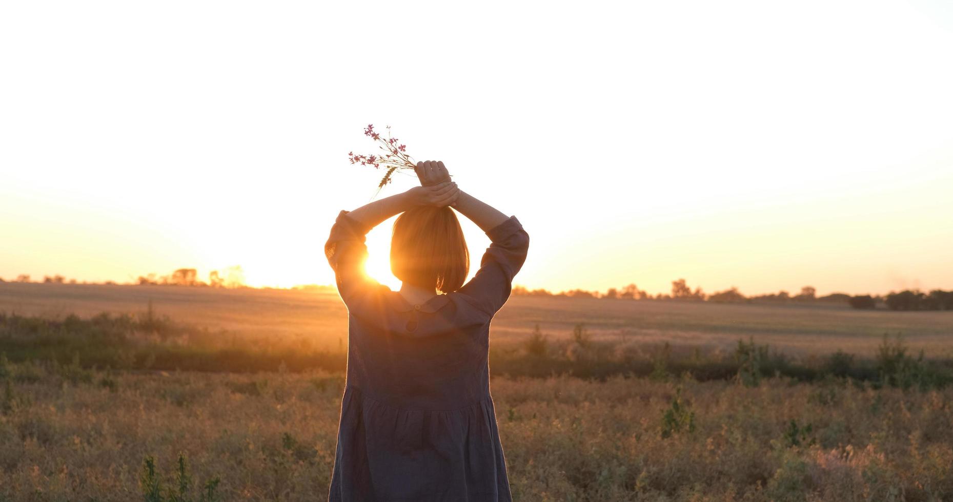 junge rothaarige frau im schönen boho-kleid, die sich während des nebligen sonnenuntergangs auf dem feld entspannt, frau im freien mit blumenstrauß in den händen foto