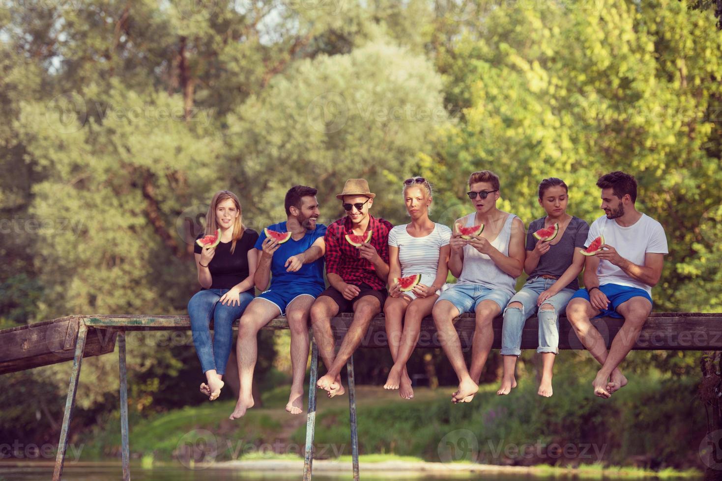 freunde, die wassermelone genießen, während sie auf der holzbrücke sitzen foto