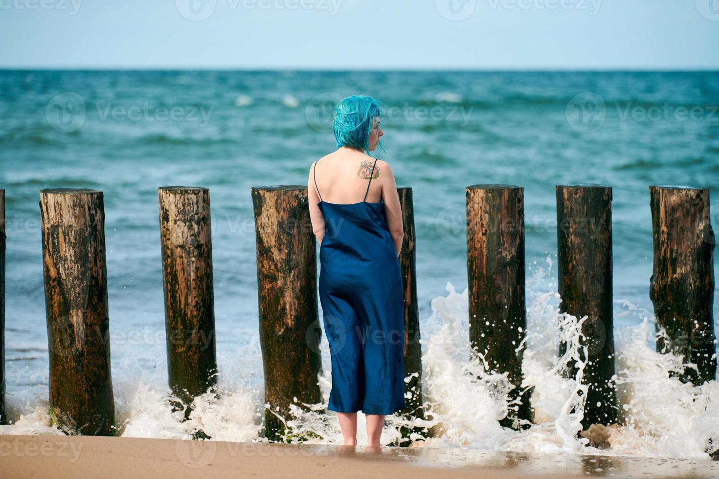 Junge blauhaarige Frau in langem dunkelblauem Kleid steht am Sandstrand und blickt auf den Meereshorizont foto