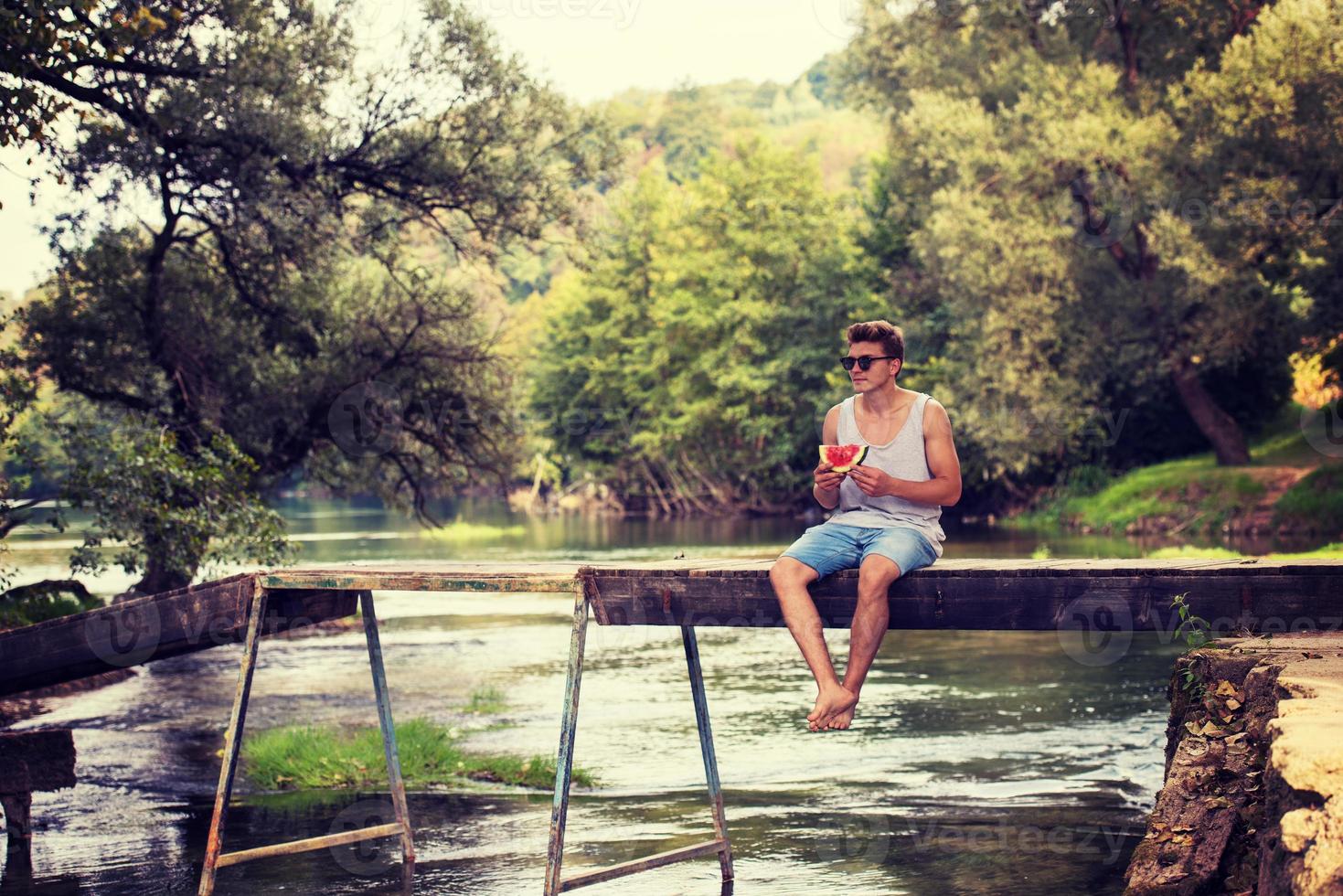 Mann, der Wassermelone genießt, während er auf der Holzbrücke sitzt foto