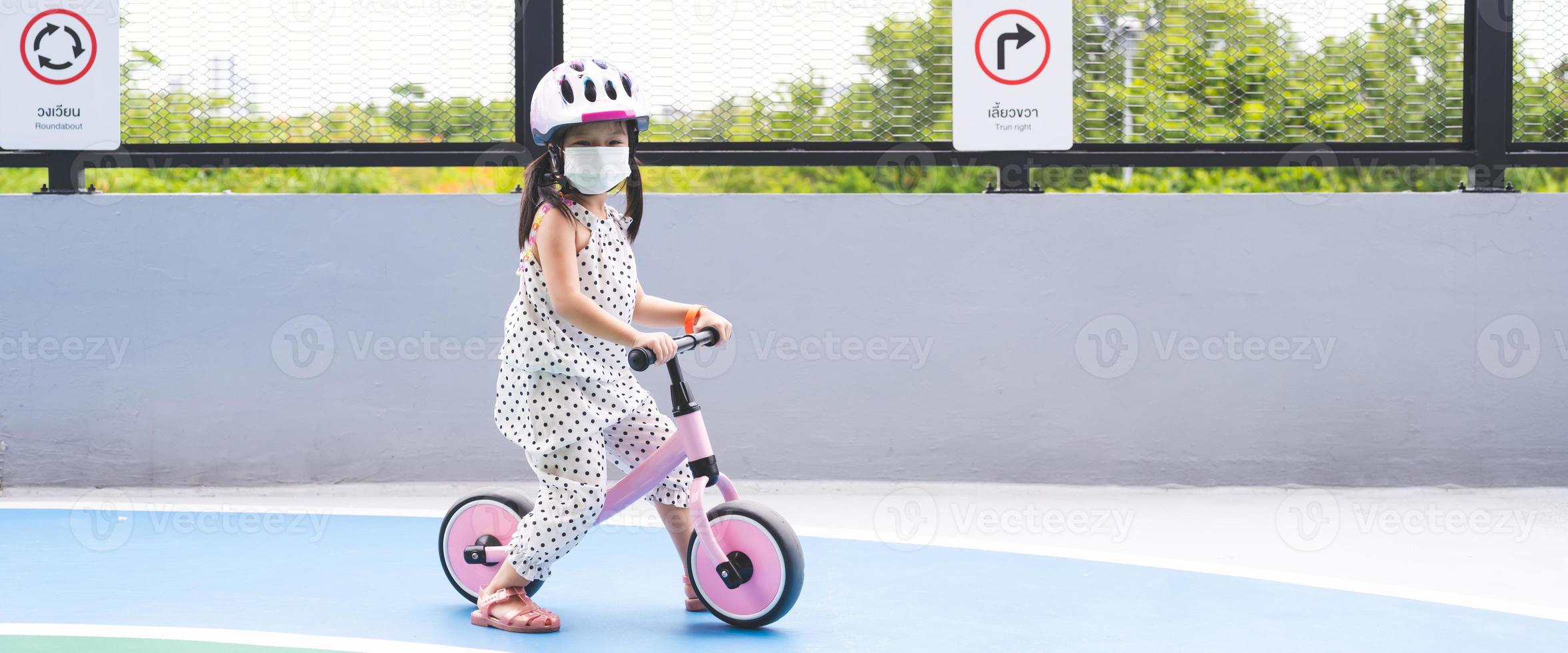süßes asiatisches Mädchen spielt auf dem Fahrrad mit ihren Beinen, die auf dem Boden gepflügt sind. Kinder tragen beim Üben Helme. hinter dem Kind ist ein Verkehrsschild in Thai und Englisch, auf dem steht: Kreisverkehr und rechts abbiegen foto