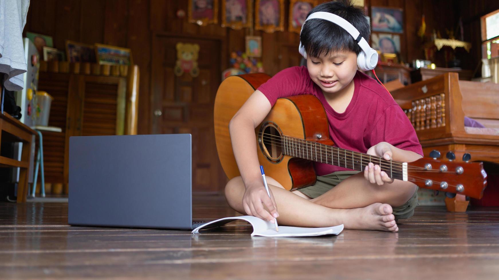 Ein süßer asiatischer Junge, der Kopfhörer trägt, ruft Videoanrufe an, lernt Musik und schreibt Songs online auf seinem Computer-Laptop mit einer akustischen Gitarre, die glücklich zu Hause ist foto