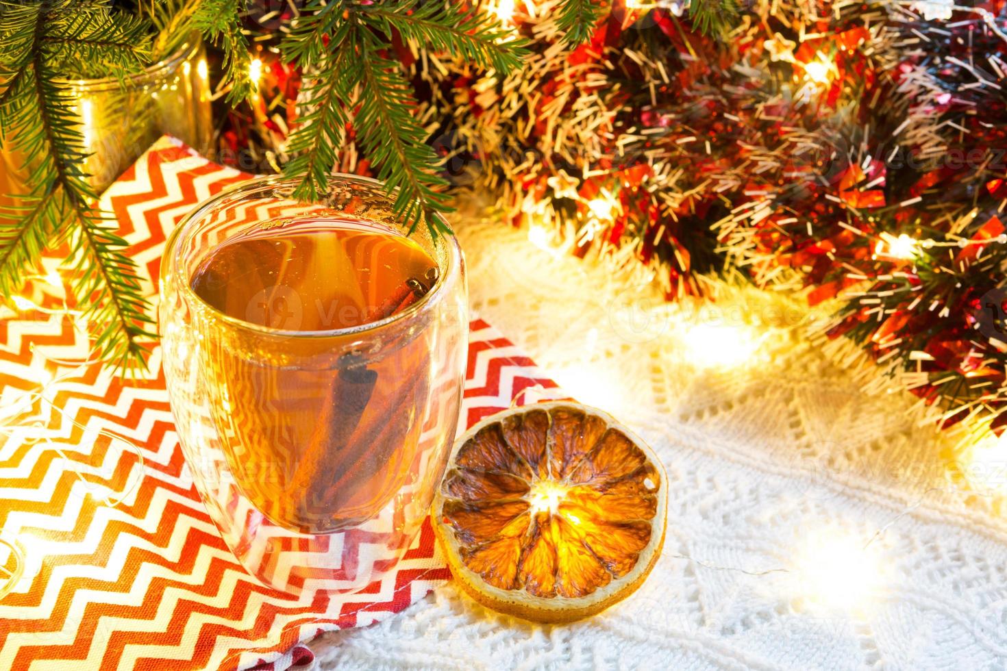 transparenter doppelwandiger glasbecher mit heißem tee und zimtstangen auf dem tisch mit weihnachtsdekor. Silvester-Atmosphäre, getrocknete Orangenscheibe, Girlande und Lametta, Fichtenzweig, gemütlich foto